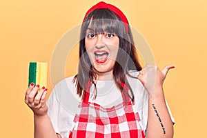 Young plus size woman wearing apron holding scourer pointing thumb up to the side smiling happy with open mouth