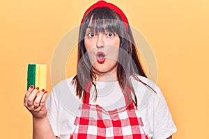 Young plus size woman wearing apron holding scourer scared and amazed with open mouth for surprise, disbelief face