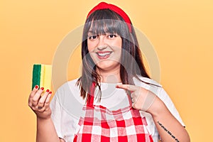 Young plus size woman wearing apron holding scourer smiling happy pointing with hand and finger