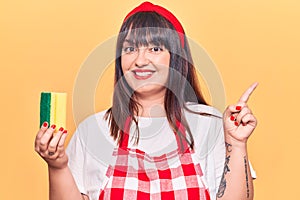 Young plus size woman wearing apron holding scourer smiling happy pointing with hand and finger to the side
