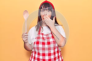 Young plus size woman wearing apron holding wooden spoon covering mouth with hand, shocked and afraid for mistake