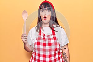 Young plus size woman wearing apron holding wooden spoon scared and amazed with open mouth for surprise, disbelief face