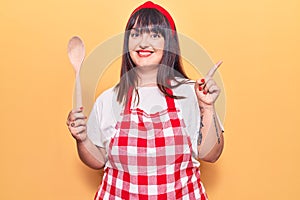 Young plus size woman wearing apron holding wooden spoon smiling happy pointing with hand and finger to the side