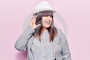 Young plus size woman wearing architect hardhat smiling with hand over ear listening and hearing to rumor or gossip