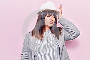 Young plus size woman wearing architect hardhat surprised with hand on head for mistake, remember error