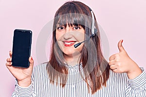 Young plus size woman wearing call center agent headset holding smartphone smiling happy and positive, thumb up doing excellent