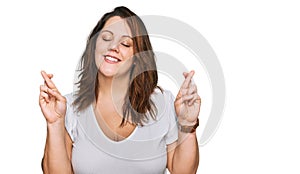 Young plus size woman wearing casual white t shirt gesturing finger crossed smiling with hope and eyes closed