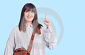 Young plus size woman wearing leather bag surprised with an idea or question pointing finger with happy face, number one