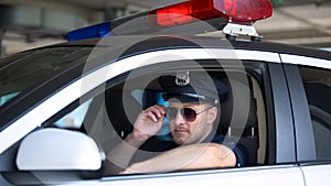 Young policeman adjusting sunglasses sitting in patrol car, responsible job