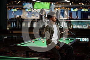 Young pool player with cue sitting on billiard table