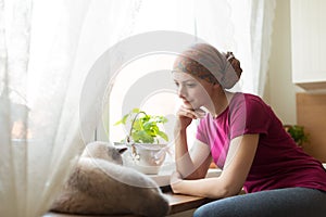 Young positive adult female cancer patient sitting in the kitchen by a window with her pet cat.