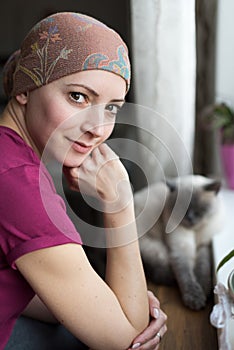 Young positive adult female cancer patient sitting in the kitchen by a window with her pet cat, smiling.