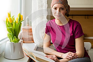 Young positive adult female cancer patient sitting in the kitchen by a window reading a book, smiling.