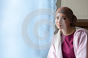 Young positive adult female cancer patient sitting in the kitchen by a window, smiling.