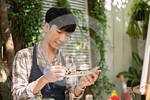 Young positive Asian man in an apron is mixing color on a palette, painting in his home garden