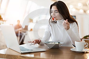 Young positive beautiful woman works in her laptop holding fork with tomatoe while eating salad in cafe during lunch time,