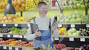 Young positive Caucasian employee posing in grocery with folder and eggplant. Portrait of smiling man in uniform working
