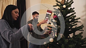 Young positive couple decorating the Christmas tree in the room before holiday. New Year and Christmas time concept