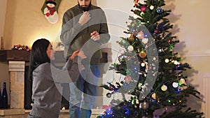 Young positive couple decorating the Christmas tree together in the room before holiday. The women giving toys to the