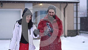 Young positive couple standing in the backyard in front of the large house under falling snow. Bearded man and