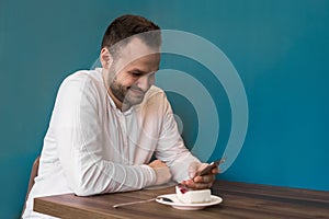 Young positive fellow European businessman in a white shirt spends time in a smartphone and smiles on a coffee break in a cafe