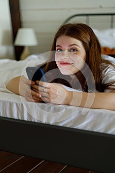 A young positive girl is lying in bed with a smartphone, watching videos, chatting with friends online