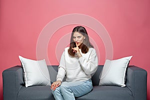 Young positive girl showing silence gesture, ask to stay quiet, keep secret about surprise sitting on cozy sofa