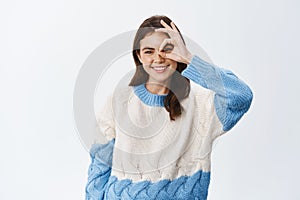 Young positive girl in sweater, showing okay sign, making alright gesture over eye and smiling, standing against white
