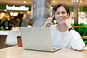 Young positive happy smiling beautiful brunette woman wearing white sweatshirt sitting at a shopping center at a table