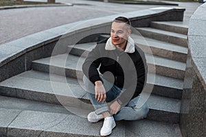 Young positive joyful man in jeans in a stylish jacket in sneakers sits and smiles on a vintage stone staircase in the city.