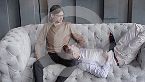 Young positive man lying on couch with laptop and talking with male partner. Caucasian gay couple resting indoors on