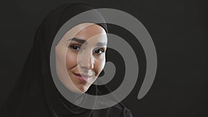 Young positive middle eastern woman in traditional headscarf smiling to camera, black studio background with empty space