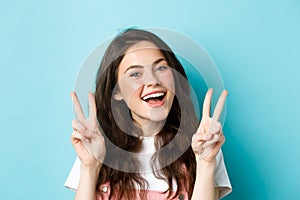 Young positive modern girl sending good vibes only, showing v-sign and smiling happy, standing against blue background