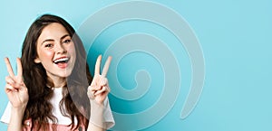 Young positive modern girl sending good vibes only, showing v-sign and smiling happy, standing against blue background