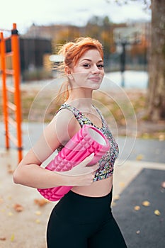 Young positive redhead sportswoman holding a foam roller in the park at autumn