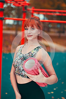 Young positive redhead sportswoman holding a foam roller in the park at vibrant autumn