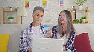 Young positive sian couple sitting at the laptop use a credit card and look into the camera