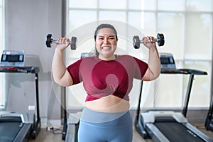 Young, Positive, Smiling, Cheerful overweight woman training with dumbbells in gym. Fat woman in sportswear doing fitness exercise