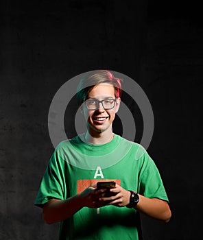 Young positive teen boy in green t-shirt and glasses standing, texting message on phone and smiling over dark background