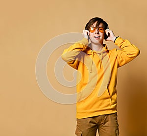 Young positive teen boy in yellow hoodie, sunglasses and headphones standing and smiling