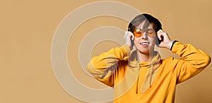 Young positive teen boy in yellow hoodie, sunglasses and headphones standing and smiling