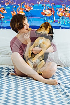 Young positive woman playing with pet dog at home interior. Funny female holding biscuit in mouth and hugging her cur dog. They