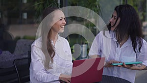 Young positive woman sitting at table with laptop greeting friend meeting in sidewalk cafe outdoors. Caucasian friends