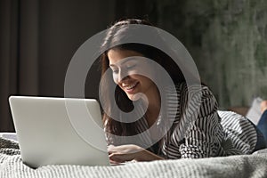 Young positive woman using computer lying on bed at home