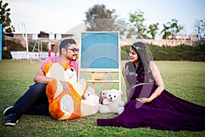 Young pregnant asian indian couple looking at each other sitting in a garden with lots teddy bear toys and blank chalkboard to