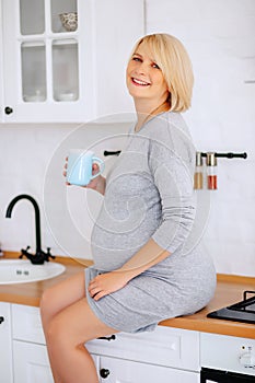 Young pregnant blonde in a gray dress sits on the kitchen table and holds a glass in his hand