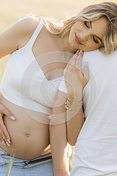 Young pregnant blonde with loose hair and naked belly, closing her eyes, clings to her husband& x27;s shoulder in nature