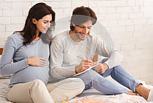 Young pregnant couple choosing baby name while sitting on bed together