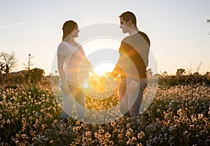 Young pregnant couple holding hands into white flowers field with the sunset and sun rays in the background