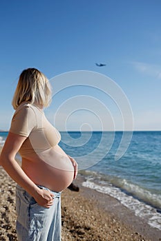Young pregnant girl by the sea looks at a flying plane